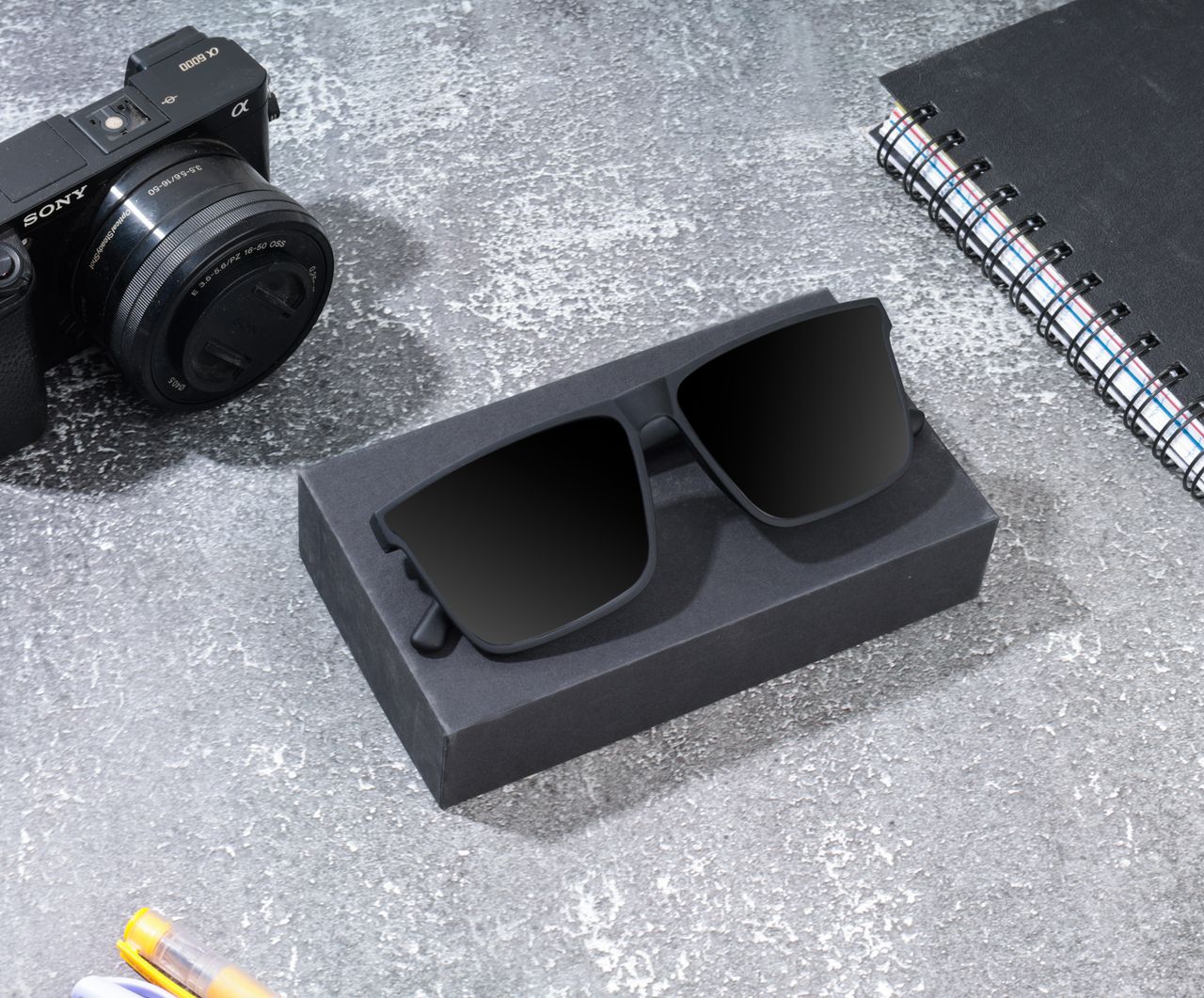 Modern sunglasses and camera on a textured desk with notebook and pen.