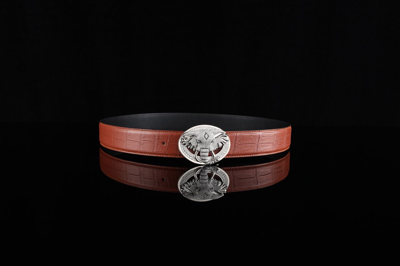 Brown leather belt with a detailed silver buckle on black reflective surface.