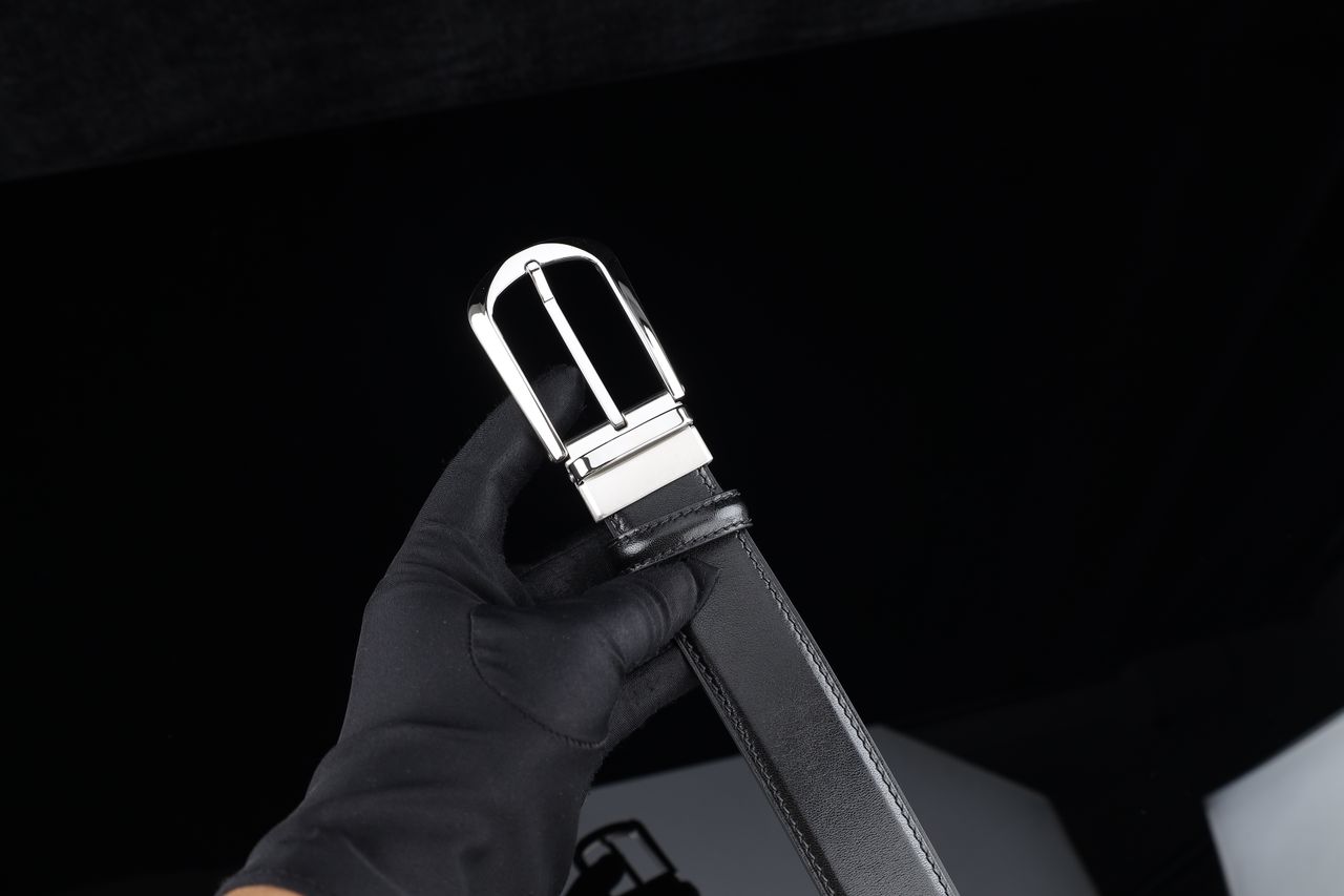 A stylish black leather belt with a silver buckle held by a gloved hand against a dark background.