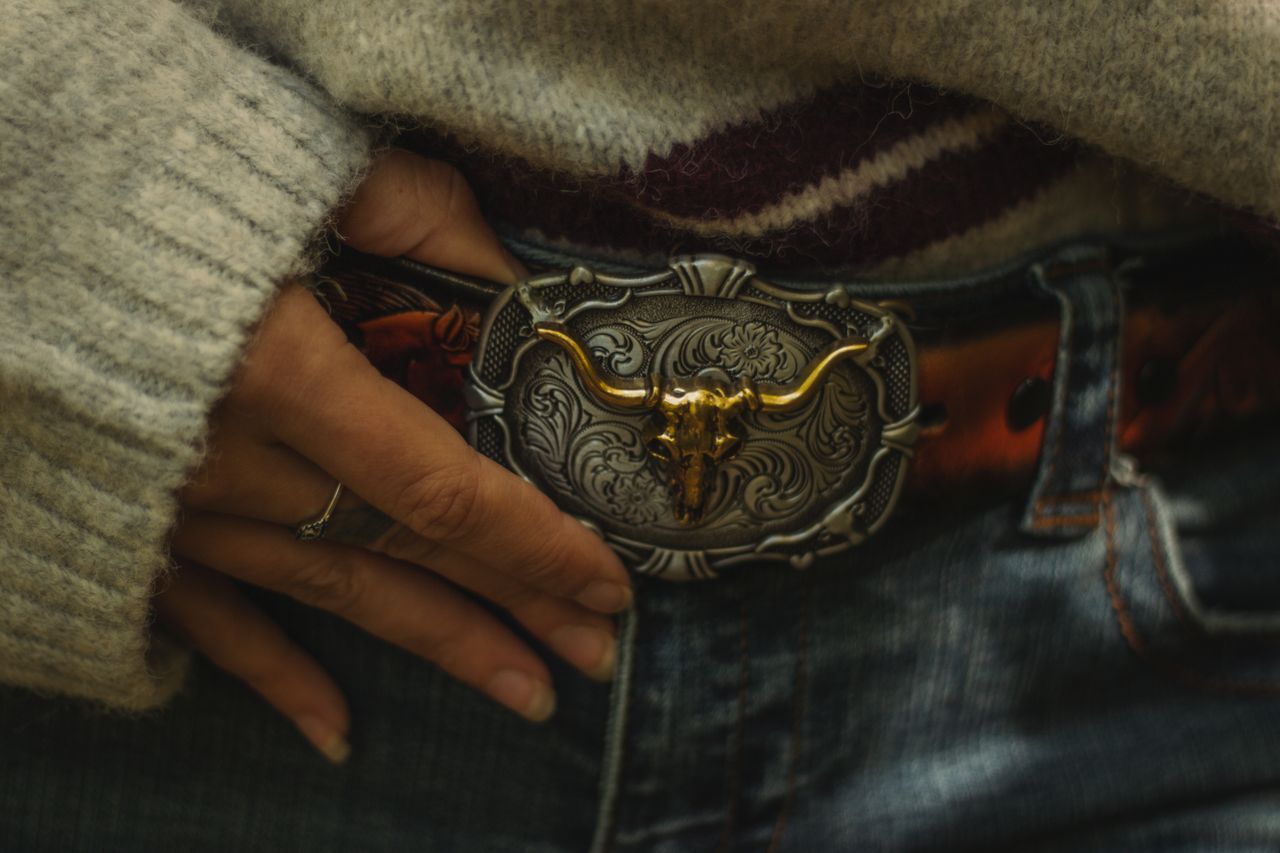 A detailed close-up of a belt buckle with intricate bull skull design, worn with denim jeans.